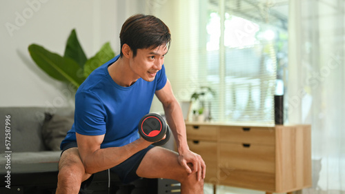 Sporty young man exercising with dumbbell in cozy home interior. Healthy lifestyle and fitness concept