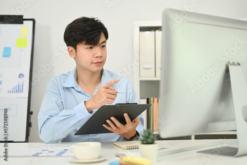 Asian man investor holding clipboard and checking online marketing on computer screen