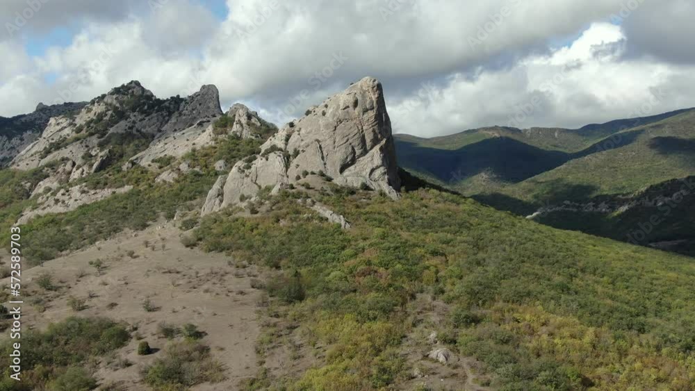 The view from the air on the cliffs among the mountains of the mountain ridge partially overgrown with forests under the blue sky with white clouds on which shadows from the clouds move. The view from the air on the cliffs among the mountains of the mountain ridge partially overgrown with forests under the blue sky with white clouds on which shadows from the clouds move.