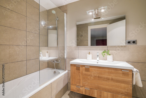 Bathroom in beautiful beige color. Vanity top sink with wooden drawers. Bathtub is fenced with protective glass from splashes. Front door is reflected in square large mirror opposite.