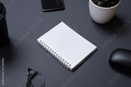 Blank notepad on black work desk witc copy space. Surounded by office supplies, phone, glasses, coffe mug, plant. Low light business composition photo