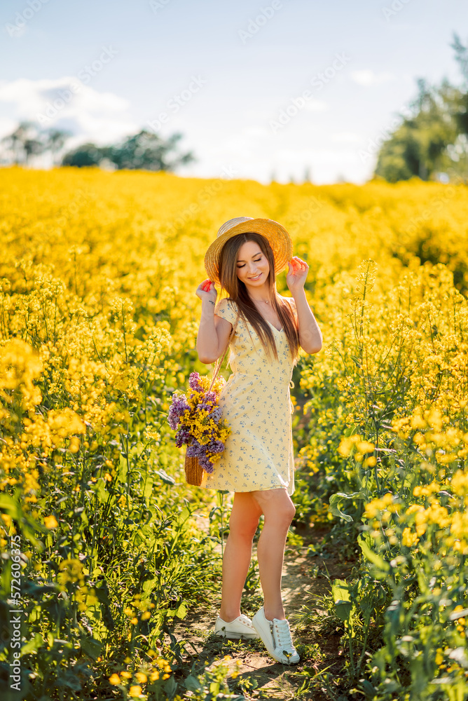 Fototapeta premium girl in a hat walks in a yellow field of rapeseed. A woman with a bouquet of flowers in her bag
