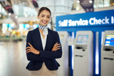 Woman, passenger assistant and arms crossed at airport by self service check in station for information, help or FAQ. Portrait of happy female services agent standing ready to assist people in travel