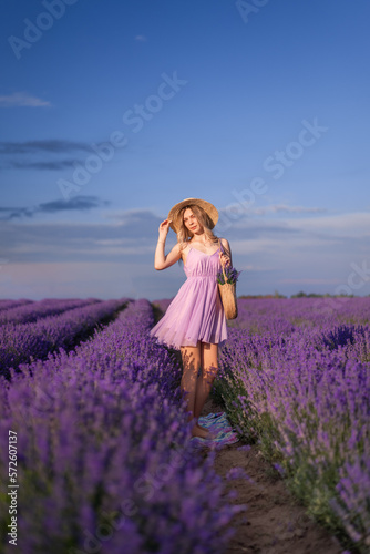 girl in a hat and a lilac dress is standing in a field of lavender. A woman with a bouquet of flowers in her purse against a beautiful sky