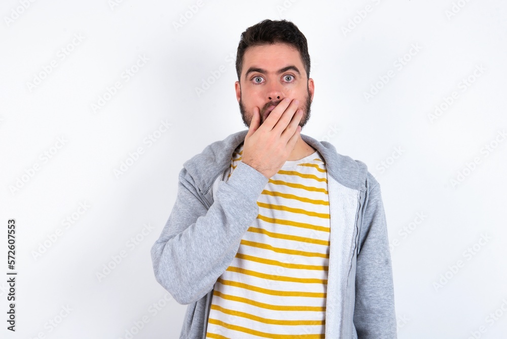 Emotional caucasian man wearing casual sportswear over white wall gasps from astonishment, covers opened mouth with palm, looks shocked at camera. Emotional caucasian man wearing casual sportswear over white wall gasps from astonishment, covers opened mouth with palm, looks shocked at camera.