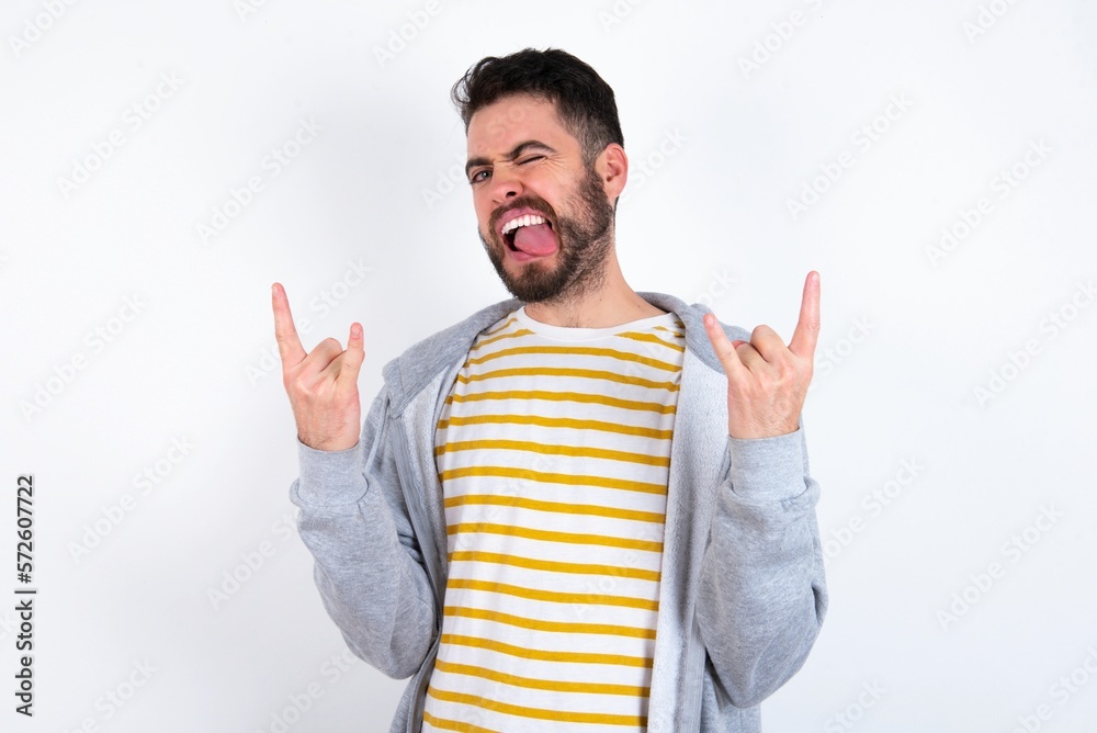 caucasian man wearing casual sportswear over white wall making rock hand gesture and showing tongue caucasian man wearing casual sportswear over white wall making rock hand gesture and showing tongue