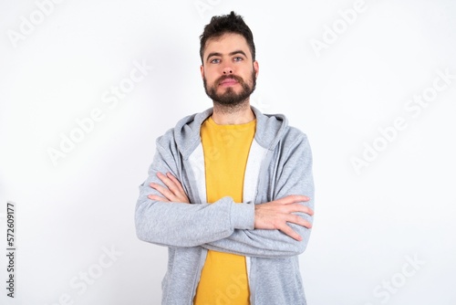 Self confident serious calm caucasian man wearing casual sportswear over white wall stands with arms folded. Shows professional vibe stands in assertive pose.