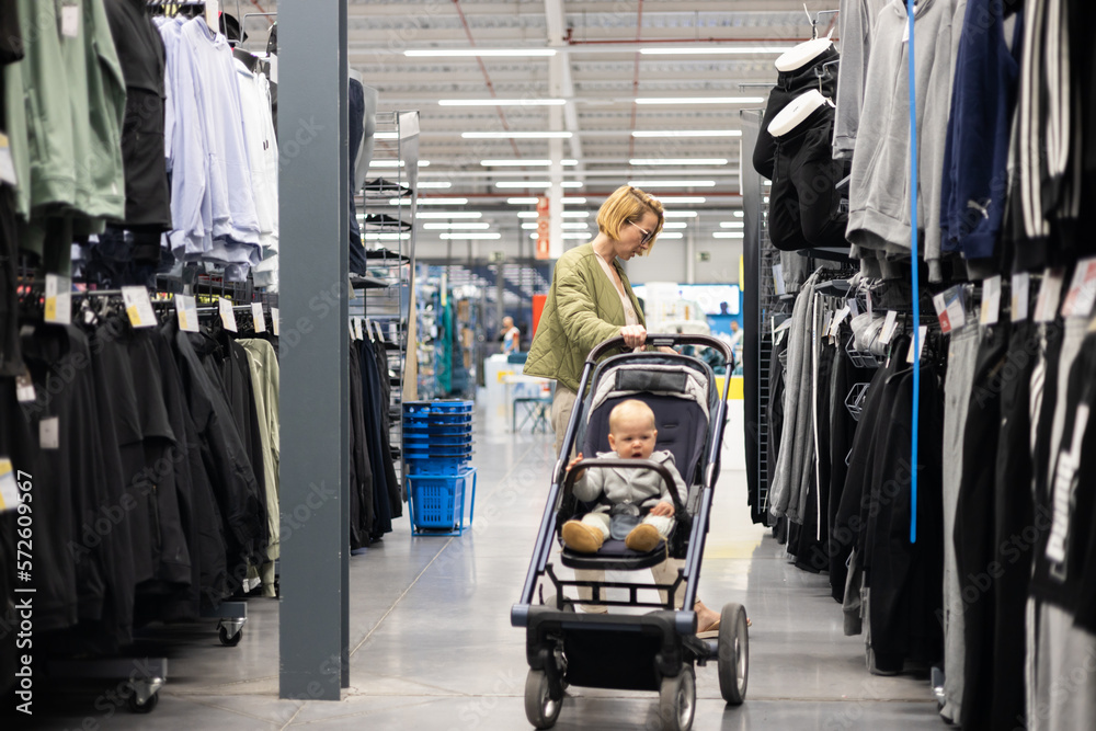 Casualy dressed mother choosing sporty shoes and clothes products in sports department of supermarket store with her infant baby boy child in stroller - obrazy, fototapety, plakaty 