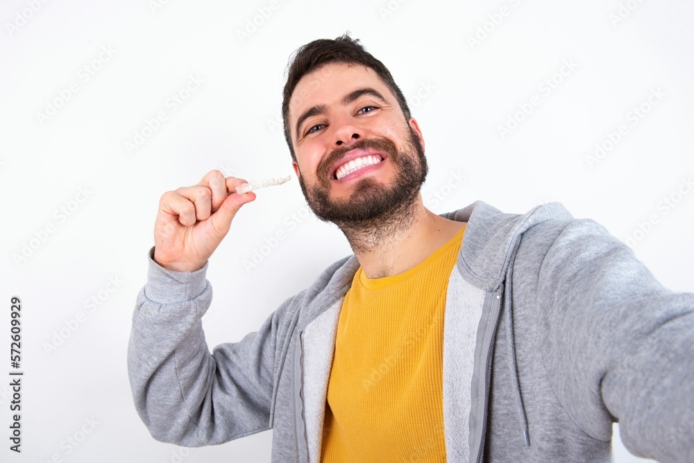 caucasian man wearing casual sportswear over white wall make selfie holding an invisible braces aligner, recommending this new treatment. Dental healthcare concept. caucasian man wearing casual sportswear over white wall make selfie holding an invisible braces aligner, recommending this new treatment. Dental healthcare concept.