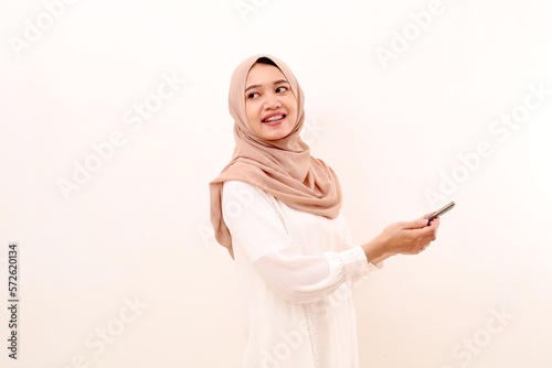 Happy young asian muslim woman looking back while holding a cell phone. Isolated on white background