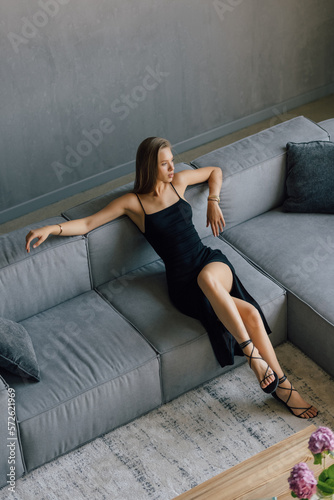 A gorgeous woman in a sexy black evening dress is lying on a black leather sofa.