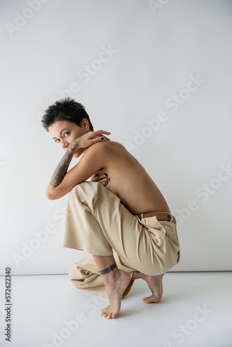 shirtless barefoot woman in beige pants looking at camera on grey background.