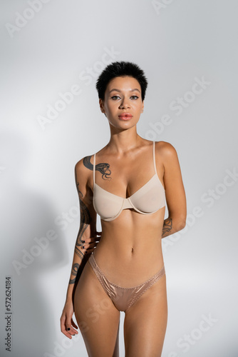 sexy tattooed woman in silk panties and bra looking at camera on grey background.