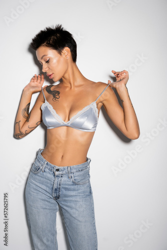 sexy woman with short brunette hair touching straps of silk bra while standing in jeans on grey background.