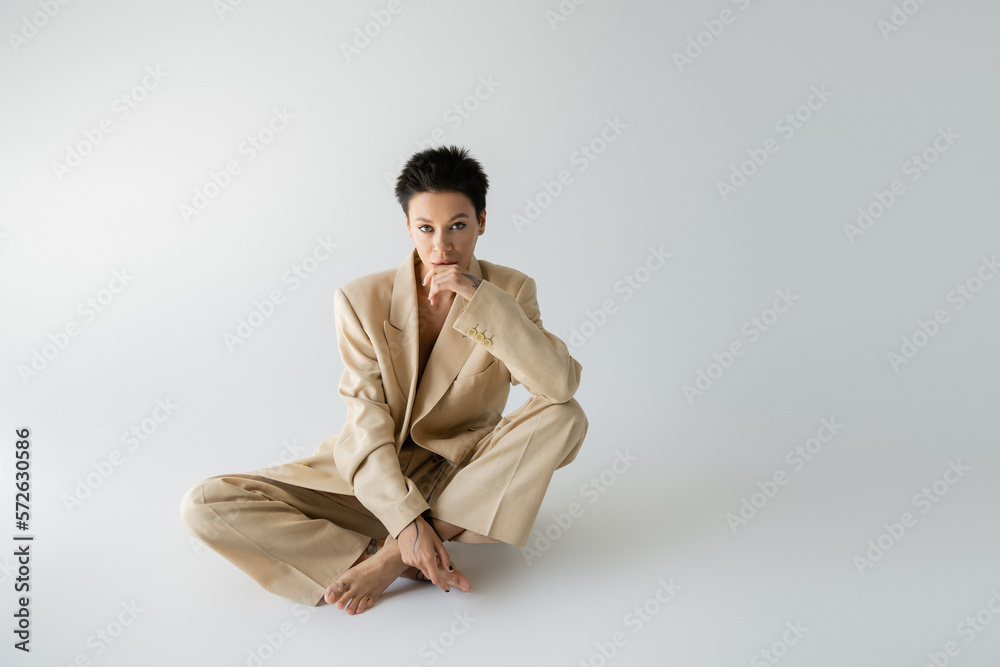 barefoot woman in beige pantsuit holding hand near face while sitting with crossed legs on grey background. barefoot woman in beige pantsuit holding hand near face while sitting with crossed legs on grey background.