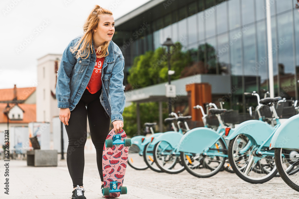Smiling young blonde girl with a jeans jacked holding a skateboard with fun print underneath getting ready to skate in the city 