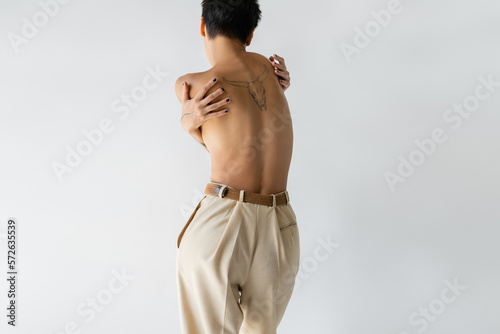 back view of tattooed shirtless woman in beige pants hugging herself isolated on grey.