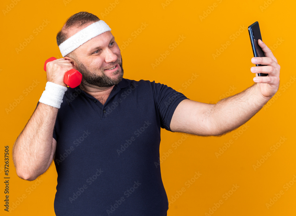 smiling adult slavic sporty man wearing headband and wristbands taking selfie holding dumbbell isolated on orange background with copy space smiling adult slavic sporty man wearing headband and wristbands taking selfie holding dumbbell isolated on orange background with copy space