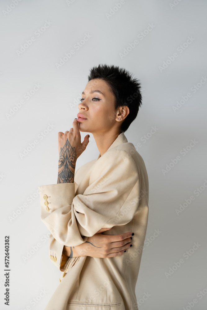 thoughtful tattooed woman in beige blazer holding hand near chin and looking away on grey background. thoughtful tattooed woman in beige blazer holding hand near chin and looking away on grey background.