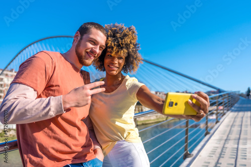 Multiracial couple through the city streets, lifestyle, selfie smiling by the river, victory gesture © unai