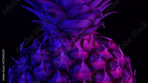 A cinematic close-up video featuring the top of a pineapple where its crown begins, rotating and illuminated by blue and purple lights in a dark style against a black background.
