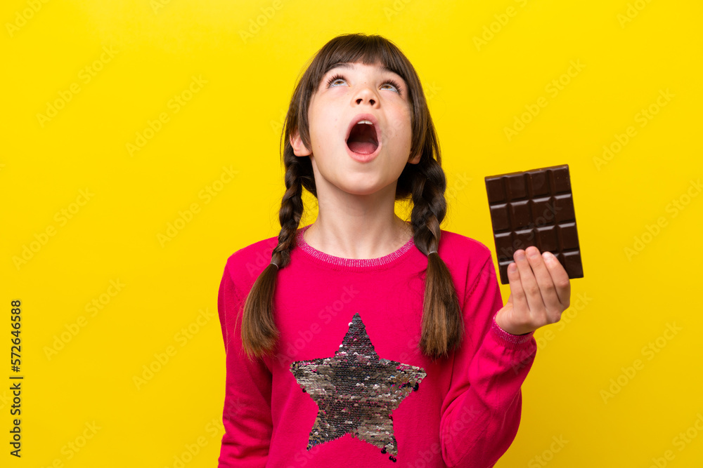 Little caucasian girl with chocolat isolated on yellow background looking up and with surprised expression Little caucasian girl with chocolat isolated on yellow background looking up and with surprised expression