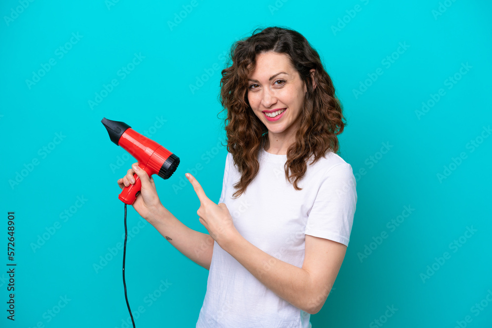 Young caucasian woman holding a hairdryer isolated on blue background and pointing it Young caucasian woman holding a hairdryer isolated on blue background and pointing it