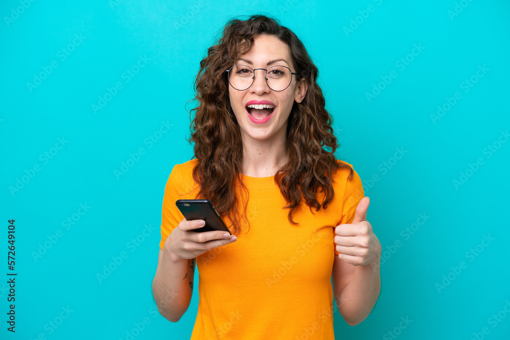 Young caucasian woman isolated on blue background using mobile phone while doing thumbs up Young caucasian woman isolated on blue background using mobile phone while doing thumbs up