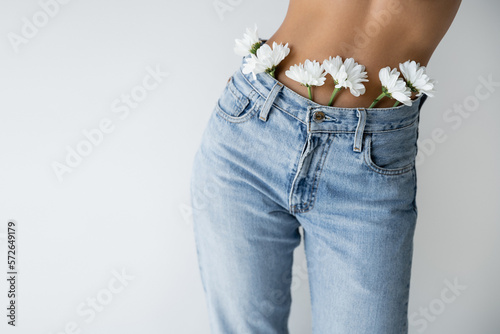 partial view of shirtless woman with white flowers in blue jeans isolated on grey.
