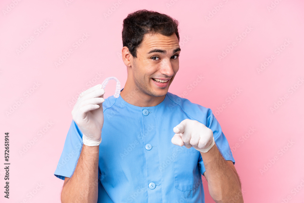 Dentist caucasian man holding invisible braces isolated on pink background surprised and pointing front