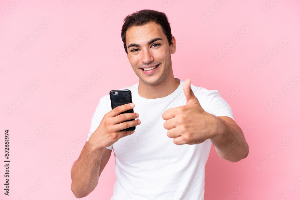 Young caucasian man isolated on pink background using mobile phone while doing thumbs up Young caucasian man isolated on pink background using mobile phone while doing thumbs up