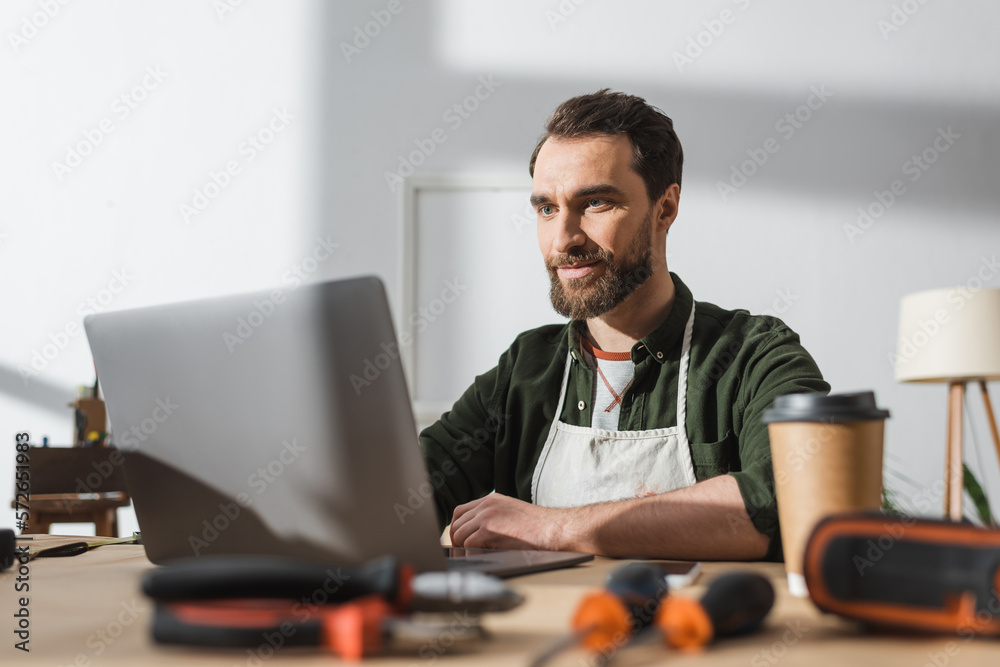 Bearded carpenter using laptop near blurred takeaway coffee and tools in workshop. Bearded carpenter using laptop near blurred takeaway coffee and tools in workshop.