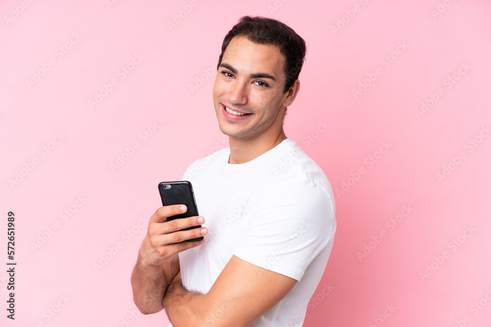 Young caucasian man isolated on pink background holding a mobile phone and with arms crossed