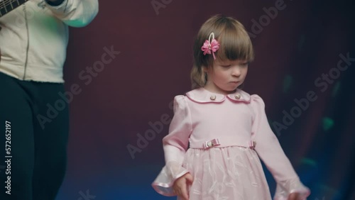 Little girl on stage in pink vintage dress, she dances, her father plays acoustic guitar. Color music is shining. Performance on stage. Early development of children