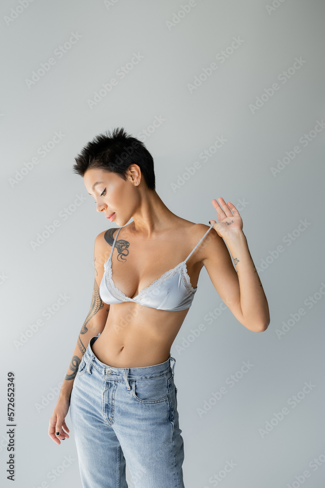 seductive brunette woman touching strap of blue bralette while posing in blue jeans isolated on grey. seductive brunette woman touching strap of blue bralette while posing in blue jeans isolated on grey.