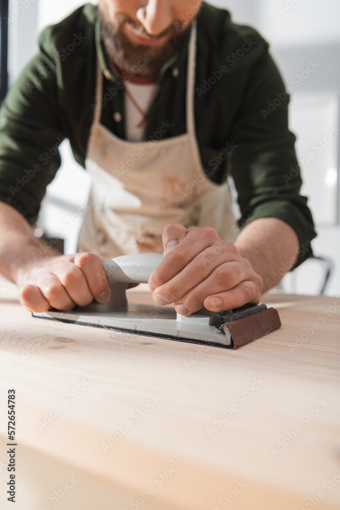 Cropped view of blurred bearded restorer sanding wooden board in workshop. Cropped view of blurred bearded restorer sanding wooden board in workshop.