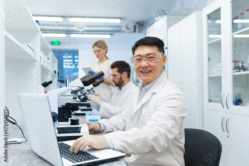 Portrait of asian lab technician, man with team group of people working in laboratory researching, man with microscope in white coat and goggles smiling and looking at camera. © Liubomir