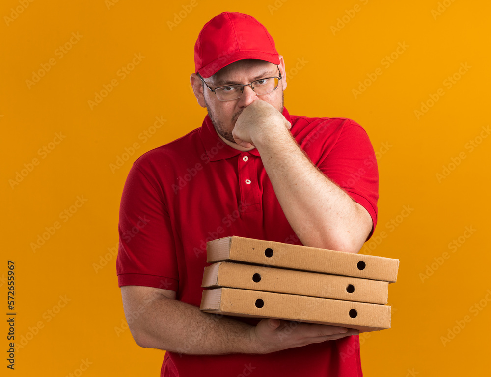thoughtful overweight young deliveryman in optical glasses holding pizza boxes and putting hand on chin isolated on orange background with copy space thoughtful overweight young deliveryman in optical glasses holding pizza boxes and putting hand on chin isolated on orange background with copy space