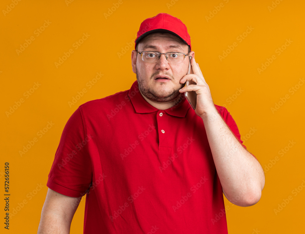 scared overweight young deliveryman in optical glasses talking on phone isolated on orange background with copy space scared overweight young deliveryman in optical glasses talking on phone isolated on orange background with copy space