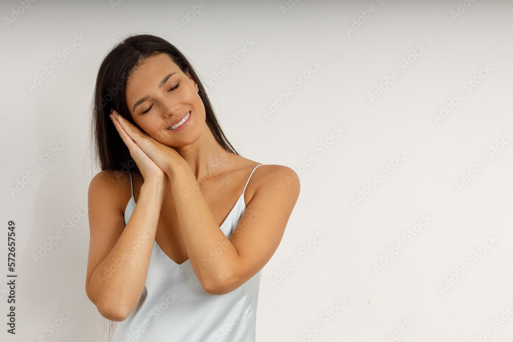 Portrait of attractive happy girl with closed eyes, holding folded hands near her face. Sleeping young brunette woman with long hair in white negligee on isolated background. Portrait of attractive happy girl with closed eyes, holding folded hands near her face. Sleeping young brunette woman with long hair in white negligee on isolated background.