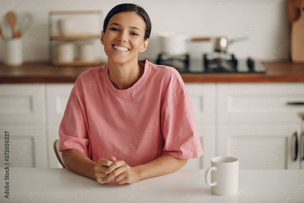 Cute young brunette woman in casual clothes looks straight into the camera. Portrait of smiling happy girl sitting at home in the kitchen at the dining table. Diet, fasting. Cute young brunette woman in casual clothes looks straight into the camera. Portrait of smiling happy girl sitting at home in the kitchen at the dining table. Diet, fasting.