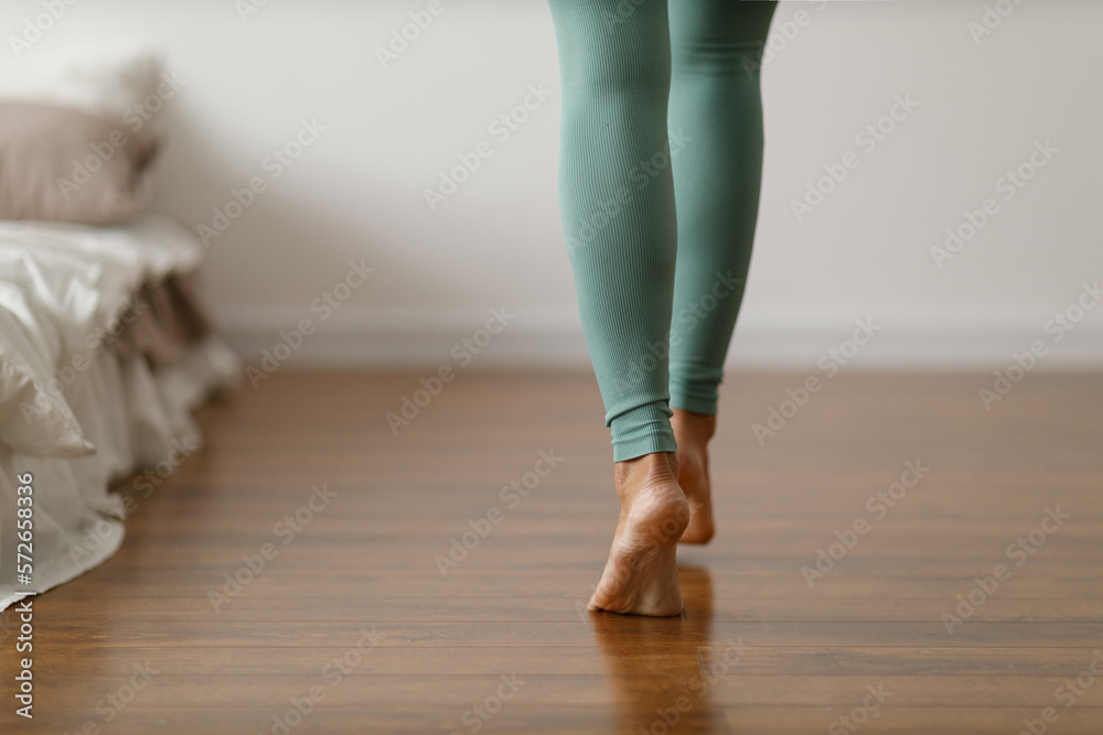 Young woman in sports leggings walks barefoot on the warm floor in the  bedroom. Close-up of cropped image of barefoot girl. Underfloor heating  system and laminated wood panels. Stock Photo