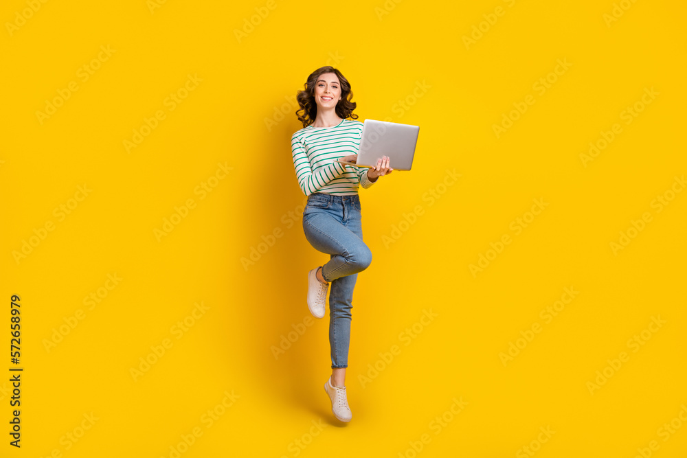 Full size photo of adorable cute nice cheerful woman dressed striped shirt jeans shoes hold laptop isolated on yellow color background Full size photo of adorable cute nice cheerful woman dressed striped shirt jeans shoes hold laptop isolated on yellow color background