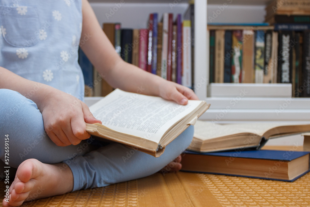 Little kid sitting in the front of bookshelf and holds few books in the hands. Concept of education