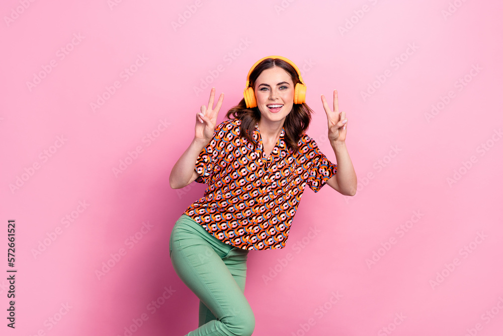 Photo of funky dancing positive girl youngster wear stylish outfit show v-sign listen headphones carefree isolated on pink color background