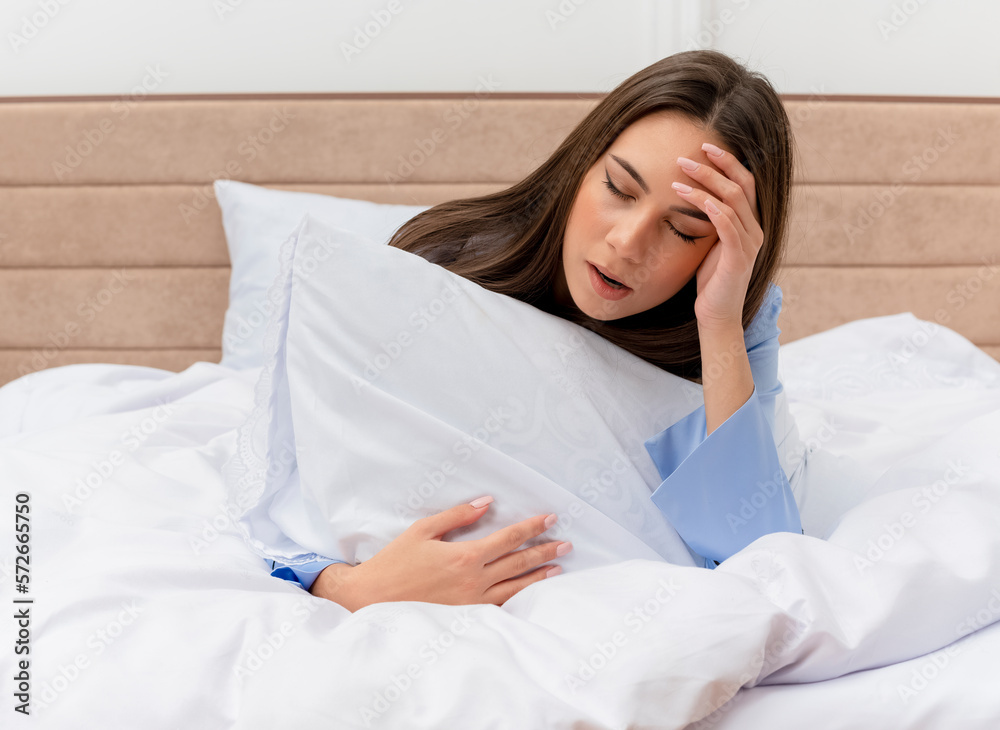 young beautiful woman in blue pajamas laying on bed with pillow yawning feeling morning fatigue in bedroom interior on light background young beautiful woman in blue pajamas laying on bed with pillow yawning feeling morning fatigue in bedroom interior on light background