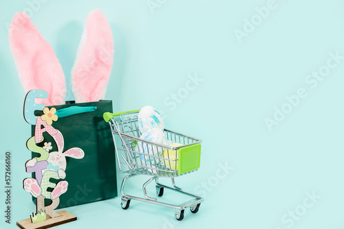 Multi-colored Easter eggs in a shopping cart, a green bag with rabbit ears. Easter shopping concept. Place for text. High quality photo