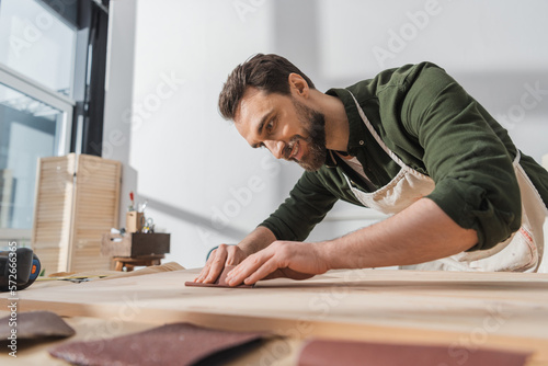 Smiling bearded workman sanding wooden board in workshop.