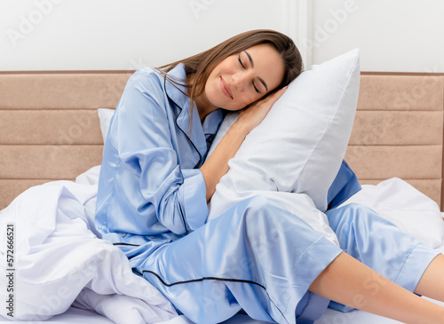 young beautiful woman in blue pajamas sitting on bed with pillow resting enjoying morning time in bedroom interior on light background © HN Works