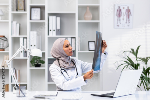 Young successful female doctor in hijab working in clinic inside medical office, muslim woman is angry and thinking x-ray picture sitting at workplace with laptop.